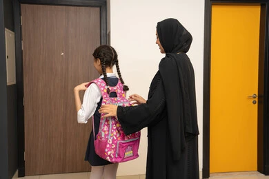 The role of parents in children's lives, preparing for the start of a new school year, perseverance and hard work to reach the highest ranks, the concept of learning in advanced schools in the United Arab Emirates, a picture from the back of a veiled Emirati Gulf Arab mother wearing a black abaya helping her daughter get ready to go to school