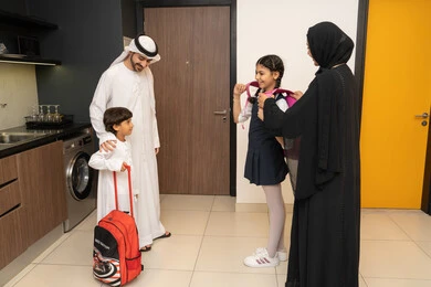 العودة إلى المدارس، أجواء صباحية عائلية داخل المنزل، رجل عربي خليجي إماراتي يرتدي الكندورة والغترة يستعد للنزول مع اولادة، ام عربيه خليجيه اماراتية ترتدي العباءه تقوم بمساعدة ابنتها فى ارتداء الحقيبه المدرسية