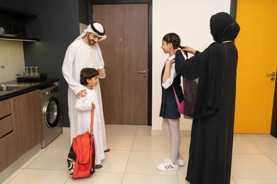 التحضير لبداية العام الدراسي الجديد، أجواء صباحية عائلية داخل المنزل، رجل عربي خليجي إماراتي يرتدي الكندورة والغترة يستعد للنزول مع اولادة، ام عربيه خليجيه اماراتية ترتدي العباءه تقوم بتعديل شعر ابنتها قبل النزول للمدرسة