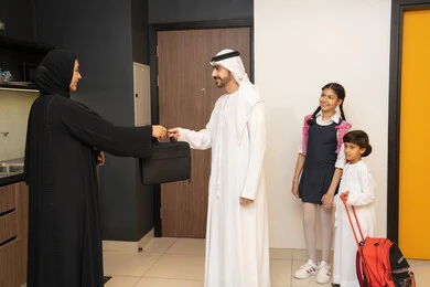 استقبال وتوديع أفراد العائلة، تواصل أسري دافئ، رجل عربي خليجي إماراتي يرتدي الكندورة والغترة يقف عند باب المنزل يودع زوجته و ياخذ حقيبة العمل، اطفال خليجيه يرتدون الزي التقليدي يستعدون للذهاب الى المدرسة ، أجواء ترحيبية داخل المنزل