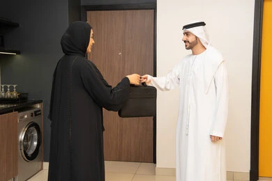 الاستعداد للذهاب إلى دوام العمل بنشاط, رجل عربي خليجي اماراتي يرتدي الزي التقليدي ياخد حقيبه العمل من زوجته, الاهتمام بالأناقة والمظهر الخارجي، سيده اماراتية تقوم بمساعدة زوجها اثناء ذهابة الى العمل