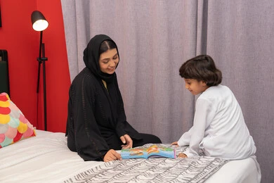 تنميه المهارات الفكريه للأبناء، تشجيع الأولاد على التعلم والتثقف، القيام بالانشطة المسلية في المنزل، سيده سعودية تقوم بقضاء وقت ممتع مع الأبناء، ام عربية خليجيه اماراتية ترتدي العباء السوداء تجلس على السرير بجانب ابنها و تقوم بقراءة القصص والحكايات قبل النوم