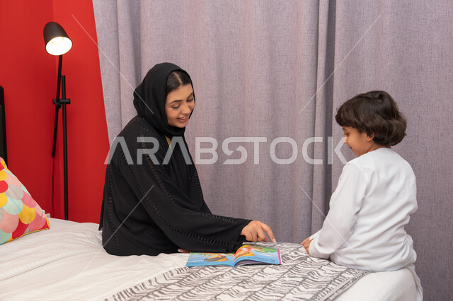 Encouraging children to learn and educate themselves, doing fun activities at home, a Saudi woman spending quality time with her children, an Emirati Gulf Arab mother wearing a black abaya sitting on the bed next to her son and reading stories and tales before bed