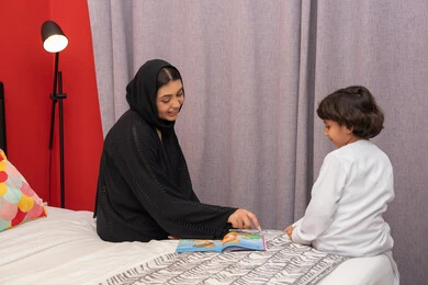 Encouraging children to learn and educate themselves, doing fun activities at home, a Saudi woman spending quality time with her children, an Emirati Gulf Arab mother wearing a black abaya sitting on the bed next to her son and reading stories and tales before bed