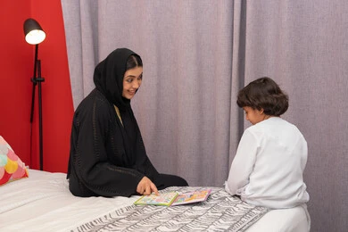 مفهوم السعاده الأسرية وإهتمام الأم بالأبناء، القيام بالانشطة المسلية في المنزل، تعليم الاولاد على التثقيف والمطالعة، سيده سعودية تقوم بقضاء وقت ممتع مع الأبناء، ام عربية خليجيه اماراتية ترتدي العباء السوداء تجلس على السرير بجانب ابنها و تقوم بقراءة القصص والحكايات قبل النوم