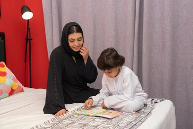 A mother's interest in her children and the concept of family happiness, doing fun activities at home, teaching children to be educated and read, a Saudi woman spending quality time with her children, an Arab Gulf Emirati mother wearing a black abaya sitting on the bed next to her son and reading stories and tales before bed