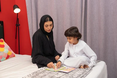 تعزيز الروابط العائلية، شغف المطالعة للكتب والروايات، تعليم الاولاد على التثقيف والمطالعة، سيده سعودية تقوم بقضاء وقت ممتع مع الأبناء، ام عربية خليجيه اماراتية ترتدي العباء السوداء تجلس على السرير بجانب ابنها و تقوم بقراءة القصص والحكايات قبل النوم