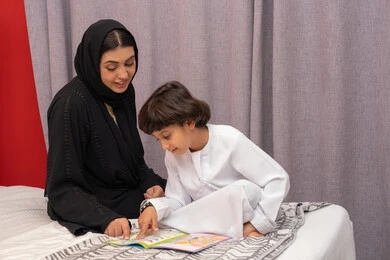 شغف المطالعة للكتب والروايات، القيام بالانشطة المسلية في المنزل، تعليم الاولاد على التثقيف والمطالعة، سيده سعودية تقوم بقضاء وقت ممتع مع الأبناء، ام عربية خليجيه اماراتية ترتدي العباء السوداء تجلس على السرير بجانب ابنها و تقوم بقراءة القصص والحكايات قبل النوم