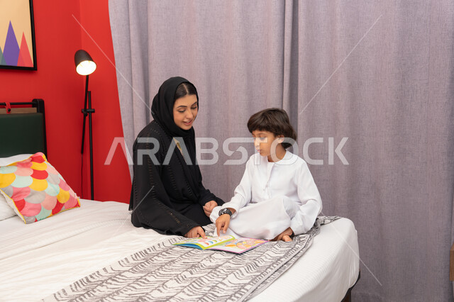 القيام بالانشطة المسلية في المنزل، شغف المطالعة للكتب والروايات، تعليم الاولاد على التثقيف والمطالعة، سيده سعودية تقوم بقضاء وقت ممتع مع الأبناء، ام عربية خليجيه اماراتية ترتدي العباء السوداء تجلس على السرير بجانب ابنها و تقوم بقراءة القصص والحكايات قبل النوم