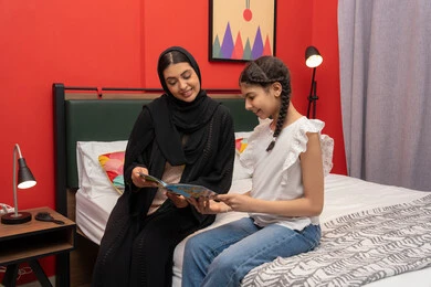 الإستمتاع بالأنشطه الترفيهية وقضاء الوقت مع الاولاد، تعزيز الروابط العائلية، ام سعودية تقوم بقضاء وقت ممتع مع الابنه، المشاركه بقراءه القصص المفيده مع الأبناء قبل النوم، بامراة عربيه خليجيه اماراتية ترتدي العباءه السوداء تجلس على السرير برفقه ابنتها تقرأ الروايات المسليه