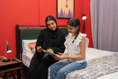 تعزيز الروابط العائلية، مفهوم التربيه القائمه على الحب والحنان، ام سعودية تقوم بقضاء وقت ممتع مع الابنه، المشاركه بقراءه القصص المفيده مع الأبناء قبل النوم، بامراة عربيه خليجيه اماراتية ترتدي العباءه السوداء تجلس على السرير برفقه ابنتها تقرأ الروايات المسليه
