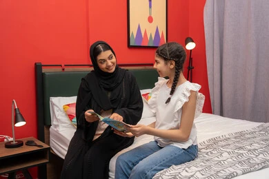 المشاركه بقراءه القصص المفيده مع الأبناء قبل النوم، مفهوم التربيه القائمه على الحب والحنان،  ام سعودية تقوم بقضاء وقت ممتع مع الابنه، بامراة عربيه خليجيه اماراتية ترتدي العباءه السوداء تجلس على السرير برفقه ابنتها تقرأ الروايات المسليه