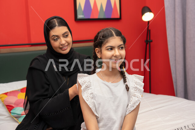 Mother's tenderness towards children, spending quality time with her daughter, the concept of parenting based on love, a smiling Emirati Gulf Arab woman wearing a black abaya sitting on the bed in the bedroom with her daughter, a Saudi woman adjusting her daughter's clothes