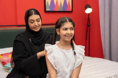 قضاء وقت ممتع مع الابنه، مفهوم التربيه القائمه على الحب، امراة عربية خليجية اماراتية مبتسمه ترتدي العباءه السوداء تجلس على السرير فى غرفه النوم برفقه ابنتها ، سيدة سعودية تقوم بتعديل ثياب طفلتها، حنان الأم على الأطفال