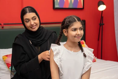 مفهوم التربيه القائمه على الحب، امراة عربية خليجية اماراتية مبتسمه ترتدي العباءه السوداء تجلس على السرير فى غرفه النوم برفقه ابنتها ، سيدة سعودية تقوم بتعديل ثياب طفلتها، قضاء وقت ممتع مع الابنه، حنان الأم على الأطفال