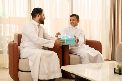 الجلوس في غرفة معيشة بتصميم كلاسيكي، التقدير والاهتمام بين الاب والابن،ايماءات البهجة والسرور، رجل عربي خليجي سعودي يرتدي الزي التقليدي  يقدم له طفله هدية باللون الأزرق، الاحتفال بالأعياد والمناسبات، الألفة والمحبة بين أفراد العائلة