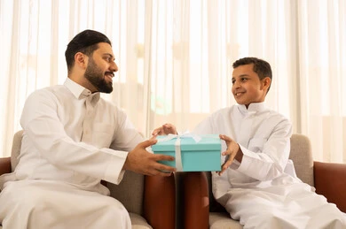 The intimacy and love among family members, celebrating holidays and occasions, gestures of joy and happiness, a Saudi Arabian Gulf man wearing traditional attire receiving a blue gift from his child, sitting in a living room with a classic design, appreciation and care between the father and son.