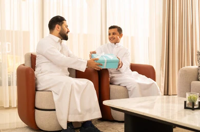 التقدير والاهتمام بين الاب والابن، الاحتفال بالأعياد والمناسبات، ايماءات البهجة والسرور، رجل عربي خليجي سعودي يرتدي الزي التقليدي  يقدم له طفله هدية باللون الأزرق، الجلوس في غرفة معيشة بتصميم كلاسيكي، الألفة والمحبة بين أفراد العائلة