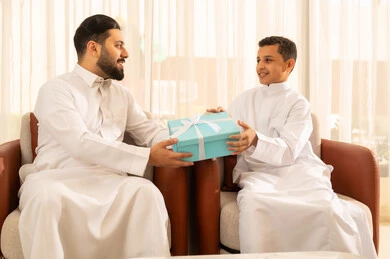 التربية الصالحة للأبناء, ايماءات البهجة والسرور، صورة مقربة لرجل عربي خليجي سعودي يرتدي الزي التقليدي  يقدم له طفله هدية باللون الأزرق،  الجلوس في غرفة معيشة بتصميم كلاسيكي، الاحتفال بالأعياد والمناسبات، الألفة والمحبة بين أفراد العائلة