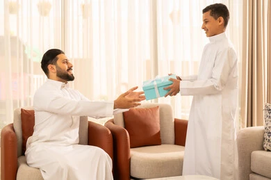 Gestures of joy and happiness, celebrating holidays and occasions, an Arab Gulf Saudi man wearing traditional attire receiving a blue gift from his child, sitting in a living room with a classic design, appreciation and care between father and son, warmth and love among family members.