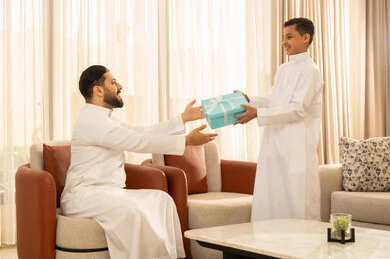 الاحتفال بالأعياد والمناسبات، ايماءات البهجة والسرور، رجل عربي خليجي سعودي يرتدي الزي التقليدي يقدم له طفله هدية باللون الأزرق، الجلوس في غرفة معيشة بتصميم كلاسيكي، التقدير والاهتمام بين الاب والابن، الألفة والمحبة بين أفراد العائلة