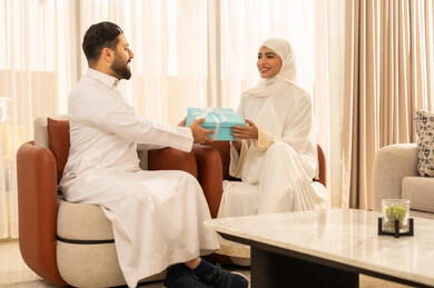 Expressing intimacy and love between spouses, giving gifts on the anniversary of the wedding day, gestures of surprise and joy, spending enjoyable family times, an Arab Gulf Saudi man wearing a white thobe presenting a special gift to his wife, the wonderful romantic atmosphere at home, a classic modern home design.