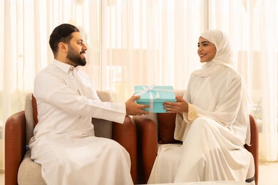 تقديم الهدايا بمناسبة ذكرى يوم الزواج، التعبير عن الألفة والمحبة بين الزوجين, إيماءات المفاجأة و السرور, قضاء الأوقات العائلية الممتعة, رجل عربي خليجي سعودي يرتدي الثوب الأبيض يقدم لزوجته هدية مميزة, الأجواء الرومنسية الرائعة في المنزل،تصميم منزل كلاسيكي عصري