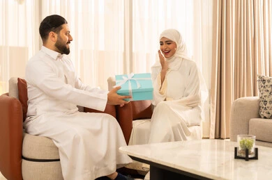 الأجواء الرومنسية الرائعة في المنزل، تقديم الهدايا بمناسبة ذكرى يوم الزواج، إيماءات المفاجأة و السرور, قضاء الأوقات العائلية الممتعة, رجل عربي خليجي سعودي يرتدي الثوب الأبيض يقدم لزوجته هدية مميزة,  التعبير عن الألفة والمحبة بين الزوجين, تصميم منزل كلاسيكي عصري