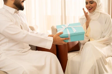 Gestures of surprise and joy, the wonderful romantic atmosphere at home, spending enjoyable family time, a close-up image of a Saudi Arabian Gulf man wearing a white thobe presenting a special gift to his wife, expressing intimacy and love between the couple, a modern classic home design.