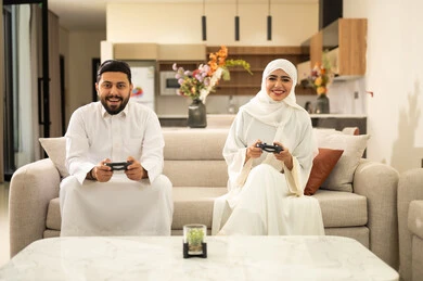مفهوم الأسرة السعيدة، قضاء وقت ممتع بلعب ألعاب الفيديو بإيماءات تدل على التركيز، زوجان عربيان خليجيان سعوديان يجلسان في غرفة المعيشة ، رجل يمسك عصا التحكم و يلعب ألعاب الفيديو مع زوجته ، المتعة و التسلية  بألعاب البلايستيشن الترفيهية