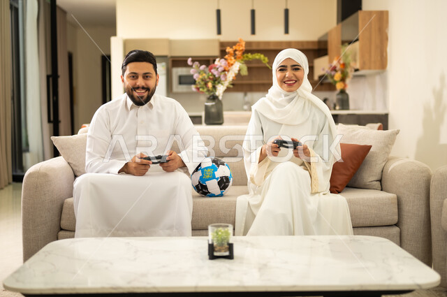 الترفيه و التسلية  بألعاب البلايستيشن الرياضية، قضاء وقت ممتع بلعب ألعاب الفيديو بإيماءات التركيز، زوجان عربيان خليجيان سعوديان يجلسان في غرفة المعيشة ، ديكور منزل عصري وكلاسيكي، رجل يمسك عصا التحكم و يلعب ألعاب الفيديو مع زوجته، مفهوم الأسرة السعيدة