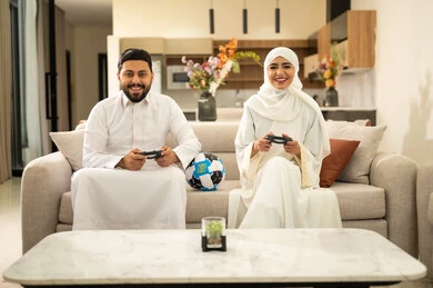 الترفيه و التسلية  بألعاب البلايستيشن الرياضية، قضاء وقت ممتع بلعب ألعاب الفيديو بإيماءات التركيز، زوجان عربيان خليجيان سعوديان يجلسان في غرفة المعيشة ، ديكور منزل عصري وكلاسيكي، رجل يمسك عصا التحكم و يلعب ألعاب الفيديو مع زوجته، مفهوم الأسرة السعيدة