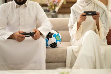 مهارة الأيدي السرية في تقنيات وألعاب البلايستيشن الرياضية، قضاء وقت ممتع بلعب ألعاب الفيديو بإيماءات التركيز، صورة مقربة لزوجان عربيان خليجيان سعوديان يجلسان في غرفة المعيشة ، ديكور منزل عصري وكلاسيكي، رجل يمسك عصا التحكم و يلعب ألعاب الفيديو مع زوجته