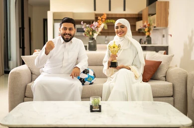 Raising a fist with expressions of victory and triumph, enthusiasm and interaction with the national team, matches and sports games, spending enjoyable leisure time with the wife, gestures of passion and encouragement for the team, a Saudi Arabian couple sitting on a comfortable sofa, the husband holding a ball in his hand while the wife holds a cup.