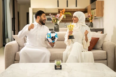 Matches and sports games, enthusiasm and interaction with the national team, raising a fist with expressions of victory and triumph, spending enjoyable leisure time with the wife, gestures of passion and encouragement for the team, an Arab Gulf couple from Saudi Arabia sitting on a comfortable sofa, the husband holding a ball in his hand and the wife holding a cup.