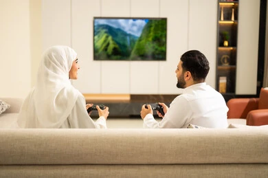 نظرات التحدي والتوعد بين اللاعبين، لعب العاب الفيديو الترفيهية ، قضاء أوقات الفراغ مع العائلة ، استخدام التكنولوجيا الحديثة، صورة من الخلف لزوجان عربيان خليجيان سعوديان يمسكان عصا التحكم، إيماءات الفوز والخسارة فى اللعبة، التفاعل والاندماج مع الالعاب الالكترونية .