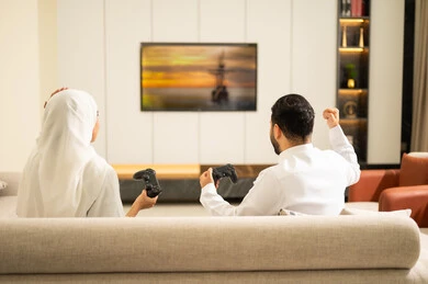 ألعاب البلايستيشن الترفيهية، لعب العاب الفيديو الترفيهية ، قضاء أوقات الفراغ مع العائلة ، استخدام التكنولوجيا الحديثة، صورة من الخلف لزوجان عربيان خليجيان سعوديان يمسكان عصا التحكم، إيماءات الفوز والخسارة فى اللعبة، التفاعل والاندماج مع الالعاب الالكترونية .