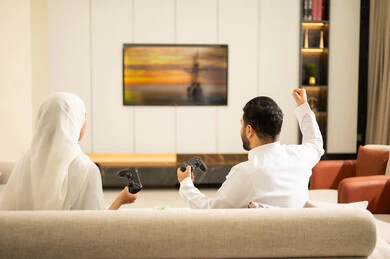 تقنيات وألعاب، ألعاب البلايستيشن الترفيهية، قضاء أوقات الفراغ مع العائلة ، استخدام التكنولوجيا الحديثة، صورة من الخلف لزوجان عربيان خليجيان سعوديان يمسكان عصا التحكم، إيماءات الفوز والخسارة فى اللعبة، التفاعل والاندماج مع الالعاب الالكترونية .