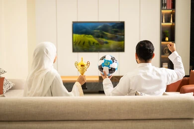 Spending enjoyable leisure time with the wife, enthusiasm and interaction with the team, raising a fist with expressions of victory and triumph, matches and sports games, gestures of passion and encouragement for the team, an Arab Gulf couple from Saudi Arabia sitting on a comfortable sofa, the husband holding a ball in his hand and the wife holding a trophy.