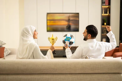 Watching and cheering at sports matches, raising a fist with expressions of victory and triumph, gestures of passion and encouragement for the team, an Arab Gulf couple from Saudi Arabia sitting on a comfortable sofa, the husband holding a ball in his hand while his wife holds a cup, enjoying leisure time with his wife, enthusiasm and interaction with the national team.