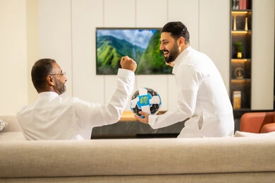 حمل الكرة باليد خلال مشاهدة المباريات، ايماءات الحماس بالفوز وتسجيل الأهداف، قضاء وقت ممتع مع الأصدقاء في المنزل، صورة من الخلف لصديقان عربيان خليجيان سعوديان يشاهدان مباراة كرة القدم في غرفة المعيشة، الجلوس في غرفة المعيشة على آريكة مريحة