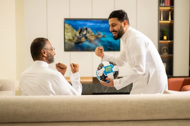 أنديه رياضية مختلفة، حمل الكرة باليد خلال مشاهدة المباريات، ايماءات الحماس بالفوز وتسجيل الأهداف، قضاء وقت ممتع مع الأصدقاء في المنزل، صورة من الخلف لصديقان عربيان خليجيان سعوديان يشاهدان مباراة كرة القدم في غرفة المعيشة، الجلوس في غرفة المعيشة على آريكة مريحة