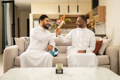 ايماءات الحماس بالفوز وتسجيل الأهداف، الشغف بمتابعة المباريات، قضاء وقت ممتع مع الأصدقاء في المنزل، صديقان عربيان خليجيان سعوديان يشاهدان مباراة كرة القدم في غرفة المعيشة، الجلوس في غرفة المعيشة على آريكة مريحة، الحماس والتفاعل مع المنتخب