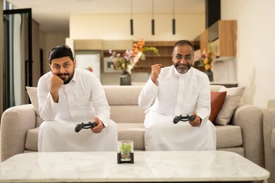 The concept of fun and entertainment, participation and competition in games, a recreational youth activity, playing recreational video games with colleagues, spending enjoyable time with friends at home, two Arab Gulf Saudi men wearing white thobes holding game controllers in their hands, sitting on a comfortable couch.