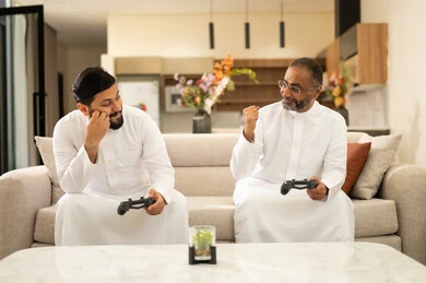 Spending enjoyable time with friends at home, the concept of fun and entertainment, sharing and competition in games, a recreational youth activity, playing entertaining video games with colleagues, two Arab Gulf Saudi men wearing white thobes holding game controllers in their hands, sitting on a comfortable sofa.