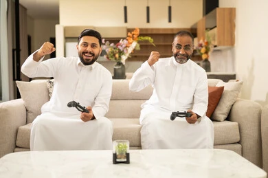 قبضة يد علامة فوز، مفهوم المتعة والتسلية، المشاركة والمنافسة بالألعاب،  نشاط شبابي ترفيهي، اللعب بألعاب الفيديو الترفيهية مع الزملاء، رجلان عربيان خليجيان سعوديان يرتديان الثوب الأبيض يمسكان بيدهما عصا التحكم، الجلوس على آريكة مريحة