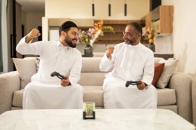 A youth recreational activity, a winning grip, the concept of fun and entertainment, participation and competition in games, playing recreational video games with colleagues, two Arab Gulf Saudi men wearing white thobes holding a game controller in their hands, sitting on a comfortable sofa.