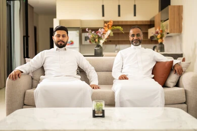 النظر إلى الكاميرا بنظرات ثقة وسرور، التسليه والترفيه الشبابي, قضاء وقت ممتع مع الأصحاب، صديقان عربيان خليجيان سعوديان يرتديان الثوب التقليدي يجلسان على آريكه مريحة باللون الابيض، الاستمتاع بالأنشطة الترفيهيه في عطلة نهاية الاسبوع