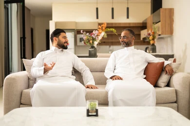 Spending enjoyable time with friends, discussion and dialogue among friends, youth entertainment and recreation, two Arab Gulf Saudi friends wearing traditional attire sitting on a comfortable white sofa, enjoying recreational activities on the weekend.