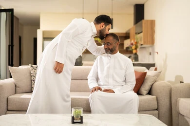 A kiss on the head is a sign of love and appreciation, spending enjoyable time with friends, discussion and dialogue among friends, youth entertainment and recreation, two Arab Gulf friends from Saudi Arabia wearing traditional attire sitting on a comfortable white sofa, enjoying recreational activities on the weekend.